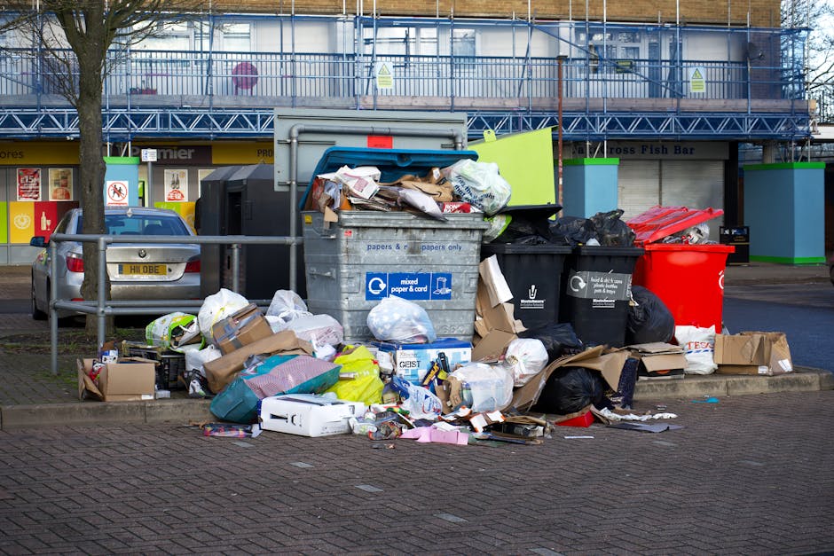 A large collection of mixed household waste and rubbish is piled on a paved sidewalk area outside a commercial building, with several black and red garbage bags, flattened cardboard boxes, and loose paper and packaging materials. The rubbish includes crumpled paper, plastic containers, and cardboard boxes, some of which are partially torn or crushed. The waste is situated next to a metal railing, with a silver car parked behind it. In the background, there are storefronts with signs, a tree on the left, and a building with a blue temporary construction barrier or scaffolding. The scene appears to be an outdoor location for rubbish disposal, possibly left for collection or an instance of excess waste. This setting highlights the need for proper waste management and the role of independent rubbish removal services such as those provided by Rubbish Removal Kentish Town, especially when dealing with non-standard or bulk waste outside regular council collections.