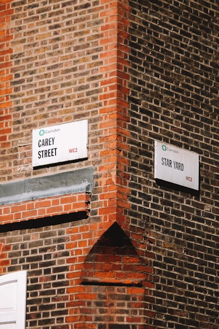 A close-up view of a corner on a brick building, showing two white street signs attached to the reddish-brown and dark brown brick wall. The left sign reads 'Carey Street WC2' and the right sign states 'Star Yard WC2', both marked with the Camden Council logo. Below the Carey Street sign, a grey metal vent box is embedded into the wall, and a horizontal brick ledge extends beneath it. The wall surfaces display a mixture of textured bricks with reddish, brown, and black tones, some bricks appearing weathered. The scene is illuminated by natural light, emphasizing the textures and contrasting colors of the bricks and signs, creating a neutral, professional visual suitable for content related to urban environment or local authority signage.
