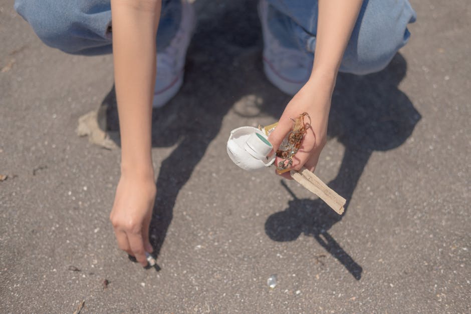 A person is crouching on a paved outdoor surface, holding a small, damaged white ceramic object with visible green paint, alongside a piece of light-colored wood and some tangled plant fibers or organic debris. The individual is dressed in casual clothing, including light blue jeans and white sneakers with red accents. The background shows the textured asphalt ground, with small scattered stones and dirt, indicating an outdoor environment. The scene appears to involve waste or rubbish, consistent with rubbish removal services, such as those provided by Rubbish Removal Kentish Town, especially in contexts of on-site clearance or independent waste disposal. Natural sunlight illuminates the scene, casting shadows that emphasize the textures of the ground and the objects held. The composition centers on the hand gripping the debris, evoking a focus on manual rubbish collection and disposal activities.