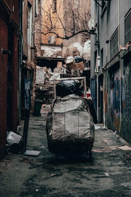 A narrow urban alleyway strewn with assorted rubbish and discarded materials, featuring a large, weathered grey duffel bag placed centrally on the uneven, dark asphalt ground. The bag appears slightly worn with visible creases and a zipper, situated in the foreground, with its handles upright. Behind the bag, there are piles of partially collapsed cardboard boxes and white plastic bags piled against a wooden fence and a brick wall, which show signs of weathering and graffiti. To the right, a dark grey metal building with barred windows and external pipes runs along the length of the alley, while on the left, an adjacent brick wall with some graffiti is visible. The background includes leafless tree branches extending over the waste heaps and surrounding buildings, under an overcast sky that provides diffuse lighting, typical of an urban environment where independent waste collection or dumping occurs. This scene aligns with private rubbish removal services operating outside standard council collections, emphasizing the importance of proper waste management and alternative disposal methods in urban settings.