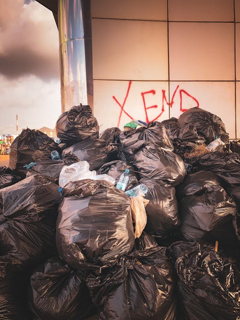 A large pile of black plastic rubbish bags stacked against a tiled wall, with some bags slightly torn or bulging, revealing miscellaneous waste. Several clear plastic water bottles are visible among the bags, some partially crushed or flattened. In the background, there is graffiti on the wall with the word 'XEND' spray-painted in red. The scene is outdoors, with a cloudy sky partially visible to the left, and the area appears to be on a paved surface, possibly a street or alleyway. The lighting suggests late afternoon or early evening, casting a warm glow on the bags and wall. The overall impression is of an unmanaged waste accumulation in an urban environment, highlighting the need for effective rubbish removal services, such as those provided by Rubbish Removal Kentish Town, for alternative waste handling or private disposal solutions.