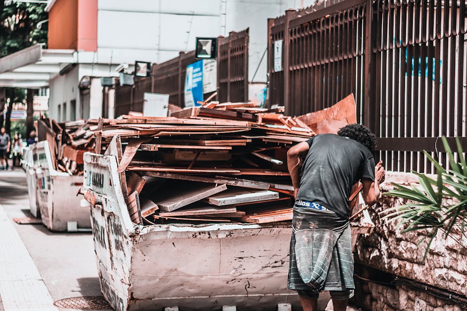 A man with curly black hair wearing a dark grey t-shirt and plaid shorts is standing on a sidewalk next to a large, white skip filled with a substantial load of construction debris, including wooden planks, tiles, and broken pieces of material. The skip, positioned near a brown metal fence, appears to be parked on a paved area adjacent to a building with glass windows and a small overhang. The wooden debris inside the skip varies in size and thickness, with some planks appearing weathered and others newer, all stacked haphazardly. In the background, a few blurred pedestrians are visible walking along the street, which features additional waste containers and urban infrastructure elements. The scene is lit by natural daylight, contributing to the clear visibility of textures such as the roughness of the wood, the smooth painted surface of the skip, and the metal fence. This visual context illustrates a typical scene of private rubbish removal or on-site clearance, reflecting the kind of waste managed by companies like Rubbish Removal Kentish Town, especially during building or renovation projects requiring specialized refuse collection.