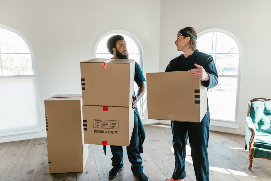 Two men are indoors in a bright room with white walls and large, arched windows allowing ample natural light. The man on the left, with a beard and curly hair, is holding two stacked cardboard boxes, each labeled with black text and red tape, and has a focused expression while looking at the other man. The man on the right, with straight hair tied back and wearing a headband, is holding a single cardboard box and gesturing with his right hand as they converse. The boxes appear to be plain, medium-sized, brown with black striped markings, and are typical of packaging used in furniture or bulk rubbish removal. In the background, there are more cardboard boxes placed on the wooden floor, and a green upholstered armchair with wooden accents is positioned near the window on the far right. The scene suggests an environment of moving, clearing, or private waste handling, in line with services offered by Rubbish Removal Kentish Town, focusing on independent collection or on-site clearance of bulky items.