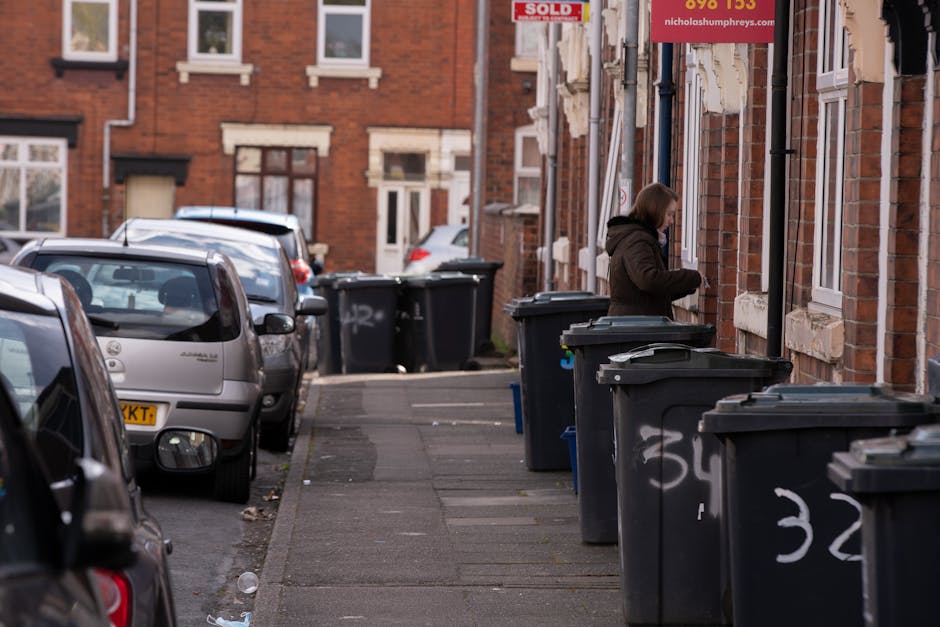 A residential street scene with a series of black wheelie bins placed along the pavement in front of red-brick terraced houses. The bins are lined up close to the houses' front walls, with some lids slightly open and graffiti markings visible on their sides. A woman, dressed in a dark coat, is standing near the bins, handling waste or rubbish, with her profile facing slightly away from the camera. In the background, additional bins are positioned further down the sidewalk, with parked cars lining the opposite side of the street. The street is paved with concrete slabs, and the environment appears to be during daylight hours with natural lighting illuminating the scene. The houses feature distinct white window frames and stone detail around the windows and doors, and the overall scene suggests a typical urban environment where private waste collection companies like Rubbish Removal Kentish Town may be involved in rubbish disposal and on-site clearance services relevant to the area.