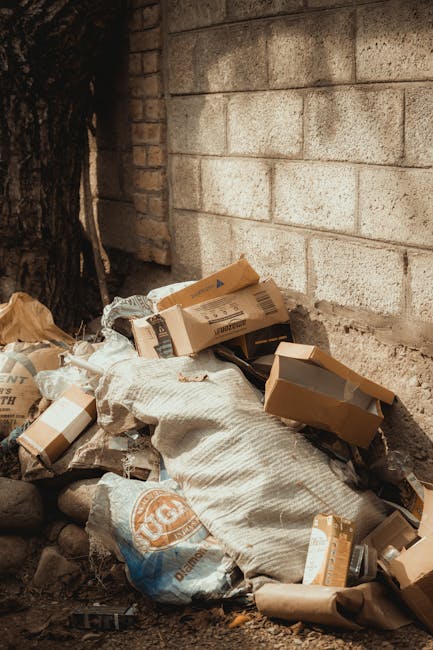 A pile of assorted waste materials and packaging bags is gathered against a rough concrete wall exterior. The waste includes cardboard boxes, some flattened and others partially intact, with visible printed labels and brown kraft textures. There are plastic and paper bags, some tied, with coarse surface textures and neutral colours like beige, brown, and white. The debris appears to be a mixture of domestic and commercial rubbish, with some boxes leaning or stacked on top of each other, partially covered by a large, crumpled cloth sack or bag. The setting is outdoors on a dirt surface with small rocks scattered around. In the background, part of a large tree trunk is visible, suggesting the waste is accumulated in a corner or alleyway. The scene indicates a typical instance of collection required for private waste handling or rubbish removal services, like those offered by Rubbish Removal Kentish Town, especially in situations involving bulk or bulky waste disposal outside of standard council collection. Natural, muted lighting emphasizes the textures and the disorderly arrangement of waste materials, highlighting the need for professional rubbish removal solutions for efficient and environmentally responsible clearance.