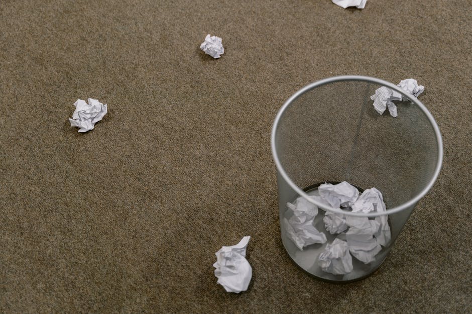 A transparent cylindrical waste bin made of clear plastic material sits on a brown textured carpeted floor, filled with crumpled sheets of white paper. Several additional crumpled paper balls are scattered across the surrounding area, some near the bin and others farther away, creating a scattered disarrayed scene. The lighting is even, highlighting the matte surface and irregular shapes of the paper debris. The environment appears to be an indoor space, possibly an office or conference room, with no other visible furnishings or objects in the frame. The presence of scattered paper and an open, empty bin suggests a waste disposal or rubbish collection scenario consistent with private or independent rubbish removal services, highlighting the need for a professional rubbish removal company like Rubbish Removal Kentish Town to manage clutter and streamline waste disposal in such settings.