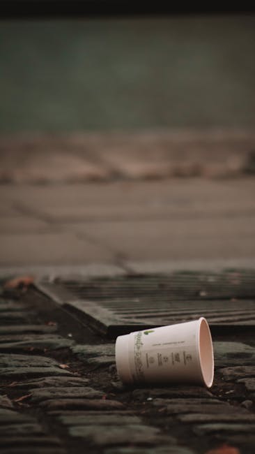 A discarded paper cup lying on a brick surface near a metal drain cover, with a wooden fence or shed in the background. The cup appears to be made of lightweight cardboard with printed text or branding on it, and it is positioned on its side, partly overlapping the rough textured bricks and the drain grate. The scene is outdoors, with natural ambient lighting creating soft shadows and highlighting the weathered appearance of the bricks and the surrounding environment. This image illustrates typical materials and conditions associated with urban waste collection and the need for professional rubbish removal services, such as those offered by Rubbish Removal Kentish Town, especially when dealing with on-site clearance or private waste handling scenarios where small litter items like paper cups are common.