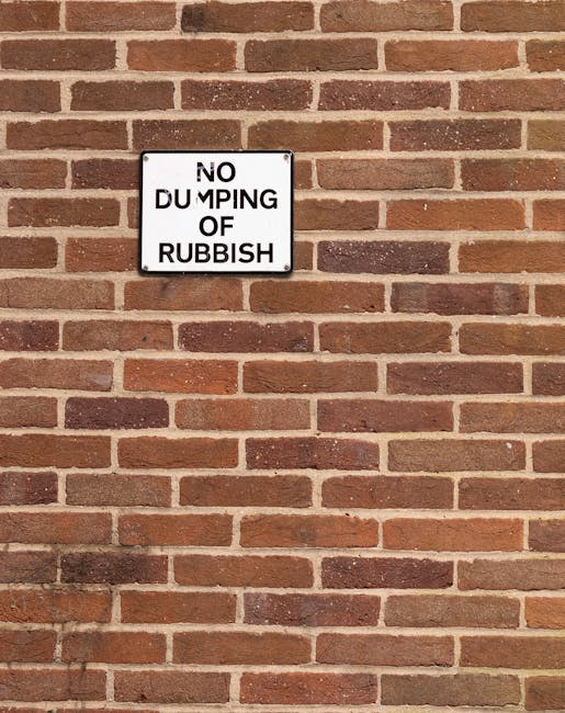 A rectangular white metal sign with black capital letters reading 'NO DUMPING OF RUBBISH' is mounted on a red brick wall. The bricks are laid in a horizontal pattern with reddish-brown and darker accent tones, and the mortar joints between the bricks are light gray. The sign is positioned roughly in the upper center of the image, slightly tilted, and appears to be securely attached. The brick wall extends beyond the edges of the photo, providing a textured, uniform background. The lighting is natural, with no shadows or highlights, creating a clear and neutral view of the scene. The overall setting suggests a private or semi-private exterior wall where rubbish dumping might occur, and the sign serves as a caution against inappropriate waste disposal, consistent with an external area that may be managed by a waste removal service like Rubbish Removal Kentish Town.