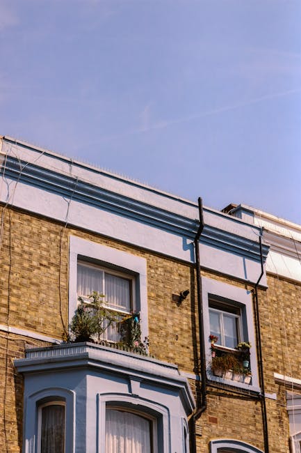 A close-up view of the upper floors of a brick residential building featuring two windows with white trim and decorative molding. The left window has a small window box with some green foliage and flowers, while the right window has a window box with a mix of plants and flowers. Black drainpipes run vertically along the facade, connecting to a gutter system at the roofline. The building's exterior displays a combination of brown brickwork and painted sections in light blue or grey. The roof has a flat surface with a metal railing, and the sky above is clear with a light blue hue, indicating a bright, sunny day. The image emphasizes the architectural details and faded painted surfaces typical of urban residential properties, with no visible rubbish or waste removal activity present.