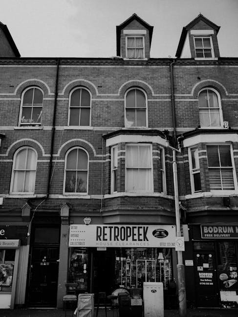 A black and white photograph depicts a three-story brick building with large, arched sash windows on the upper floors, some of which have curtains inside. The building has a prominent bay window on the second floor, with a row of smaller windows on the top floor, set within dormer structures with pitched roofs. The ground floor features commercial units; on the left, there is a store with a partially visible sign, and in the center, a window displaying the sign 'RETROPEEK HOUSE CLEARANCE' with bold lettering and an image of a car, indicating a house clearance service. The storefronts have glass windows and doors, with various items and posters visible inside. In front of the building, there are several litter bins and small traffic barriers on a paved sidewalk. Adjacent shops and signage are also visible, including one with the name 'BODRUM', suggesting a food or delivery service. The environment appears to be in an urban setting, suitable for private waste collection or rubbish removal services, aligning with alternative waste handling options.