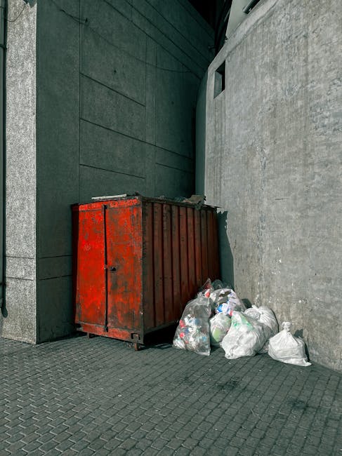 A bright red, weathered metal skip with visible rust and peeling paint is positioned in an outdoor urban setting, nestled between a dark, textured wall on the left and a light grey concrete wall on the right. The skip is ample in size, with a closed top and vertical ribbing along its sides, indicating it is designed for bulky waste collection. Adjacent to the skip, on the paved ground, are several tightly tied white plastic bags filled with rubbish, some of which are slightly bulging and leaning against the front of the skip. The surface beneath the bags has a pattern of small interlocking stones or tiles. The background environment appears to be part of a service or back alley, characterized by minimal natural light, with shadows and diffuse brightness suggesting an overcast sky or shaded area. The placement and condition of the skip and rubbish bags imply a temporary or on-site waste collection setup, reflective of private disposal or alternative waste management methods outside standard council services, aligning with independent rubbish removal practices typical of companies like Rubbish Removal Kentish Town.