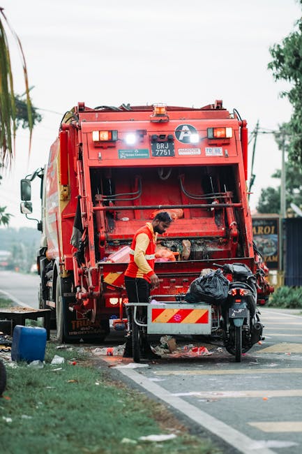 A waste collection worker in a high-visibility red and yellow uniform is operating a large red rubbish truck parked on the side of the road. The worker is focused on the rear loading area, where they are managing waste items, with a black plastic bag and some debris visible nearby. The truck, branded with safety and identification labels, features a large rear compartment with metal components and hydraulic mechanisms for waste compaction. The scene is set outdoors during daytime, with a partly cloudy sky overhead and greenery, including a tree and grass, to the left side of the image. In the background, a street with overhead power lines, traffic signs, and some commercial signage can be seen. The overall environment suggests an urban or suburban area where local waste collection services are being carried out, aligning with the themes of private or alternative rubbish removal methods that a company like Rubbish Removal Kentish Town offers, especially for on-site waste management and disposal tasks.