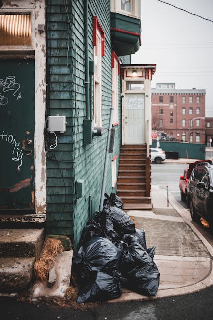 A residential building with a green wooden exterior featuring horizontal siding, situated on a street with parked cars. In the foreground, several black plastic rubbish bags are stacked against the building's wall near the base of a small set of wooden stairs leading up to a white door. The door is surrounded by a small overhang with red and white striped trim, and a white extension above it. To the left of the bags, there are some concrete steps leading to a door with graffiti markings. The environment appears to be an urban area, with a background of brick buildings and power lines visible in the overcast sky. The scene suggests a private rubbish collection or on-site waste management, as part of a rubbish removal service, with the waste ready for clearance by a local provider like Rubbish Removal Kentish Town. The setting emphasizes the typical clutter and waste accumulation outside a residential property awaiting disposal.